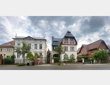 Zwei historische Wohnhäuser an einer Straße, links ein weißes Gebäude mit aufwändiger Stuckverzierung und rechts ein Fachwerkhaus in Rot und Weiß mit Erker und hohen Fenstern. Im Hintergrund bewölkter Himmel, vorne Bäume und ein Zaun.