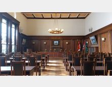 Ein historischer Versammlungsraum mit hölzernen Wänden und einem Parkettboden. Der Raum enthält zwei Reihen von Holzstühlen mit gepolsterten Lehnen, große bunte Glasfenster auf der linken Seite, ein Gemälde und mehrere Flaggen an der Rückwand sowie eine zentrale Deckenleuchte.