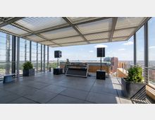 Rooftop terrace with a slatted metal canopy, glass and metal railings, and large floor tiles overlooking a city skyline. A DJ booth with speakers and cables is set up near the edge, with planter boxes containing shrubs at the corners.