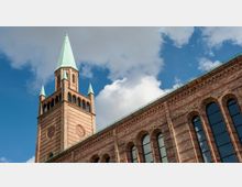 Backstein-Kirchturm mit grüner Spitze und zwei kleinen seitlichen Türmchen, rechts angeschlossen ein Gebäude mit Rundbogenfenstern; blauer Himmel mit weißen Wolken im Hintergrund.