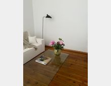 Wohnzimmer mit Holzboden, einem weißen Sofa, einer Stehlampe mit schwarzem Lampenschirm und einem Glastisch, auf dem eine Vase mit rosa Blumen und ein Magazin liegen. Der Hintergrund zeigt eine weiße Wand.