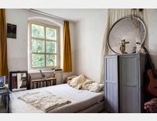 Ein schlicht eingerichtetes Schlafzimmer mit einem weißen Bett auf einer Matratze, einem grauen Schrank, gelben Vorhängen vor einem großen Fenster mit Blick auf Bäume, und einer Gitarre, die an der Wand lehnt. Bücher und Dekorationsobjekte, wie ein Kreuz und eine Vase, sind ebenfalls im Raum verteilt.