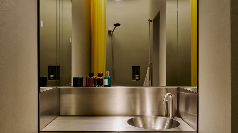 Schmales, modernes Badezimmer mit Edelstahl-Waschtisch und rundem eingelassenem Becken, flankiert von großen Spiegeln an beiden Seiten. Im Hintergrund sind eine Dusche, gelbe Vorhänge und runde Deckenöffnungen mit Beleuchtung zu sehen.