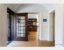 Flur in einem Wohnraum mit hellen Wänden, Holzboden und einer geöffneten Holztür mit Glaseinsätzen, die in einen weiteren Raum führt, der mit einem Holzregal und Büchern eingerichtet ist. Links ein weiterer Durchgang und rechts eine weiße Zimmertür.