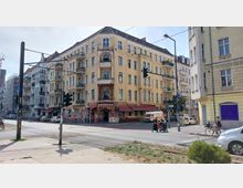 Ecke eines gelblichen Altbau-Wohnhauses mit mehreren Stockwerken und Balkonen, im Erdgeschoss befinden sich Geschäfte und ein Restaurant mit der Aufschrift "Antalya". Kreuzung mit Ampeln, Gehwegen und parkenden Autos, im Vordergrund Schienen der Straßenbahn.