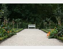 Eine gerade, kiesbedeckte Gartenallee führt zu einer weißen Holzbank, die vor dichten Bäumen steht. Zu beiden Seiten der Allee wachsen niedrig geschnittene Beete mit Blumen und Gemüse sowie junge Bäume.