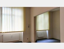 Ein leerer Innenraum mit hellbeigen Wänden und zwei großen Fenstern, die von vertikalen, gelblichen Lamellenjalousien bedeckt sind. Unter den Fenstern befinden sich weiße Heizkörper, der Boden ist mit einem dunkelgrauen Teppich ausgelegt.