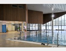 Innenansicht eines Schwimmbads mit einem rechteckigen Becken, umgeben von Startblöcken. Die Architektur zeigt Holzwände, eine hohe Decke und große Glasfenster, die Licht hereinlassen und einen Blick auf schneebedeckte Außenbäume ermöglichen.