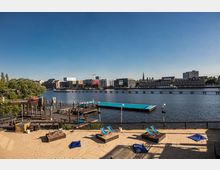 Ein Stadtstrand mit Sand, Liegestühlen und blauen Sitzsäcken im Vordergrund, angrenzend an hölzerne Stege am Wasser. Im Hintergrund sichtbar ist ein Fluss, darüber eine Fußgängerbrücke, sowie moderne Gebäude und ältere Backsteinbauten am gegenüberliegenden Ufer.