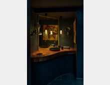 Dunkles Badezimmer mit einem geschwungenen Waschtisch aus Holz, einem runden Waschbecken und schwarzen Wandfliesen. Im Hintergrund sind ein großer Spiegel, hängende Lampen mit warmem Licht und ein Gemälde mit Blumenmotiv sichtbar.