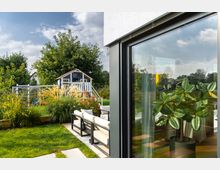 Ein moderner Garten mit einer Terrasse, die mit Sitzmöbeln ausgestattet ist, und einem kleinen, weißen Spielhaus im Hintergrund, umgeben von Blumenbeeten und Ziergräsern. Im Vordergrund spiegelt sich ein Gummibaum in einer großen Glasfront eines Hauses.