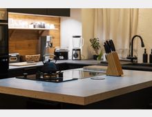 Moderne Küche mit einer Kücheninsel aus hellem Stein, darauf ein schwarzer Wasserkocher auf einem Gaskochfeld und ein Holzblock mit Messern. Im Hintergrund eine Arbeitsplatte mit Toaster, Mixer, Pflanzen und einer Spüle mit schwarzer Armatur sowie hölzernen Wandelementen.