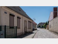 Schmale gepflasterte Dorfstraße mit einfachen Wohnhäusern mit roten Ziegeldächern und geschlossenen Fensterläden. Im Vordergrund ist ein niedriger Zaun sichtbar, im Hintergrund stehen weitere Häuser mit Vorgärten.