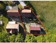 Luftaufnahme eines Gehöfts mit vier Gebäuden, umgeben von Wiesen und Bäumen. Die Grundstücksgrenzen sind durch eine rote Linie markiert, und die Gebäude haben rote Ziegeldächer sowie begrünte Außenbereiche.