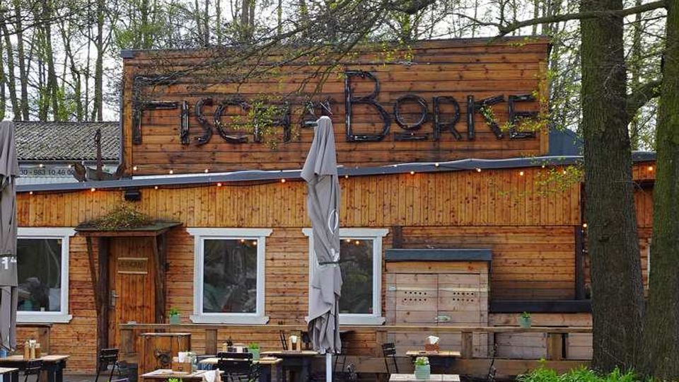 Holzgebäude mit der Aufschrift „Fischer Borke“, umgeben von Bäumen. Davor befindet sich ein Außenbereich mit Tischen, Stühlen, Sonnenschirmen und kleinen Pflanzen.