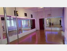 Ein Tanzstudio mit glattem Holzboden, mehreren vertikalen Tanzstangen und großen Wandspiegeln, die von farbigen LED-Lichtern eingerahmt sind. Im Hintergrund sind lila Vorhänge, rote Vorhänge und dekorative Wandbilder sichtbar.