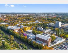 Luftaufnahme eines Industrie- und Gewerbegebiets in einer städtischen Umgebung, umgeben von vielen Bäumen und Wohngebäuden. Im Vordergrund sind mehrere Gebäude mit unterschiedlichen Fassaden, darunter ein gelbes und ein Ziegelgebäude, sowie Gleisanlagen und ein Parkplatz sichtbar.