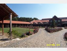 Hof mit gepflastertem Weg, umgeben von Gebäuden mit roten Ziegeldächern und Holzelementen. Im Vordergrund gepflegte Blumenbeete und ein zentraler Rasenbereich, im Hintergrund ein kleiner Wald.