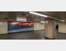 Unterführung mit gefliesten Wänden und einer farbigen Wandgestaltung, die aus orangefarbenen, blauen und grauen Flächen besteht. Der Raum ist mit Deckenleuchten beleuchtet, und ein einzelner Mann geht in der rechten Bildhälfte durch die Szene.