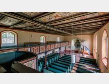 Innenansicht einer historischen Kirche mit bemalter Holzdecke, Empore mit verzierten Paneelen und grünen Holzbänken. Im Hintergrund befinden sich ein Altar mit einem verzierten Rahmen und eine Kanzel in blau-weißen Farbtönen, flankiert von bunten Glasfenstern.