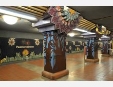 U-Bahn-Station mit kunstvoll gestalteten Säulen, die mit baumähnlichen Motiven bemalt sind, sowie Wandmosaiken mit floralen Mustern und der Aufschrift "Paulsternstraße". Der Boden besteht aus roten Fliesen, und die Decke ist mit Leuchten ausgestattet.