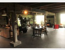 Ein offener Wohnraum mit rustikalem Design, sichtbaren Holzbalken und Betonboden. Der Raum enthält eine offene Küche, einen Esstisch mit Stühlen, Sitzmöbel, große Pflanzen sowie einen Kamin im Hintergrund. Große Fenster lassen Tageslicht herein.