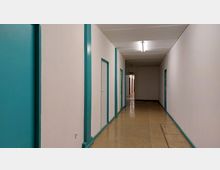 Langer, schlichter Flur mit beigen Wänden, türkisfarbenen Türrahmen und Fußleisten sowie mehreren, links angeordneten Türen. Der Boden besteht aus abgenutztem Linoleum, die Decke ist weiß mit einer langen Neonröhre.