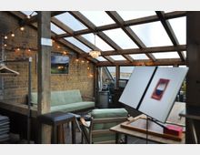 Eine gemütlich eingerichtete Veranda mit Glasdach und Holzbalken. Sichtbar sind eine Backsteinwand, eine grüne Couch, Sessel, eine Staffelei mit einem Buch, eine Hängelampe und Lichterketten, die den Raum dekorieren.