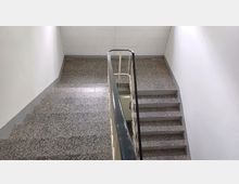 Treppenhaus eines Gebäudes mit grauem Terrazzo-Boden, weißen Wänden und einem schwarzen Geländer. Die Treppe führt in entgegengesetzte Richtungen nach oben und unten.