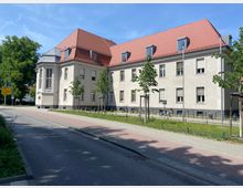 Großes, zweistöckiges Gebäude mit hellbeiger Fassade und rotem Satteldach, flankiert von jungen, neu gepflanzten Bäumen. Im Vordergrund verläuft eine Straße mit abgetrenntem Radweg; links sind Verkehrsschilder erkennbar, und im Hintergrund befinden sich weitere Bäume.