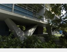 Moderne Wohnanlage mit überdachten Balkonen auf stilisierten V-förmigen Stützen, umgeben von Bäumen und Grünflächen. Im Vordergrund ist dichter Buschbewuchs, im Hintergrund sind eine Straße und Fahrzeuge sichtbar.