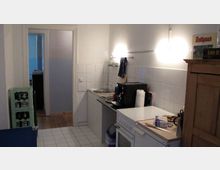 Kleine Küche mit weiß gefliesten Wänden und Boden. Zu sehen sind eine Spüle, ein Herd, eine Kaffeemaschine, ein Kühlschrank sowie Regale und Getränkekisten im Raum.