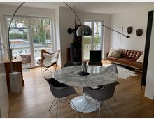 Wohnzimmer mit modernem Design: im Vordergrund ein runder Marmortisch mit grauen Stühlen, im Hintergrund ein braunes Ledersofa, eine Wandleuchte und zwei Masken als Wanddekoration. Große Fenster mit Blick auf einen Balkon und eine Außenumgebung mit Bäumen, Holzböden und skandinavischen Akzenten ergänzen den Raum.