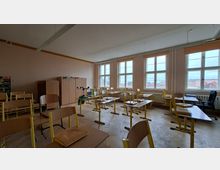 Klassenzimmer mit mehreren Schülertischen und -stühlen, teils umgedreht auf den Tischen, sowie Schränken an der Rückwand. Große, mehrteilige Fenster mit Heizkörpern darunter lassen Tageslicht herein; Holzboden und lange Deckenleuchten prägen den Raum.