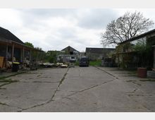 Hofbereich mit gepflastertem Untergrund, umgeben von mehreren Gebäuden mit teils wellblechgedeckten Dächern, links steht ein überdachter Bereich mit gelagerten Gegenständen. In der Mitte ein geparkter Kombiwagen und ein Anhänger, im Hintergrund Grünflächen und blühende Bäume.