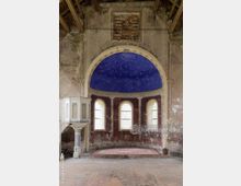 Innenraum einer verlassenen Kirche mit beschädigter Wandgestaltung. Der halbkreisförmige Altarbereich besitzt eine Kuppeldecke in Blau mit Sternen, umgeben von verwitterten Wänden und drei hohen Fenstern; links steht eine Kanzel.