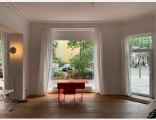 Ein heller Raum mit großer Fensterfront, durch die ein städtischer Gehweg mit Bäumen und parkenden Autos sichtbar ist. Im Vordergrund stehen ein roter Tisch mit einer Blumenvase und eine Stehlampe; der Boden ist aus Holz, und weiße Vorhänge rahmen die Fenster ein.