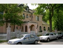 Eine rosafarbene Villa im klassizistischen Stil mit weißen Fensterläden und Rundbogenfenstern, umgeben von einem weißen Holzzaun und dichten Bäumen. Im Vordergrund parken mehrere Autos entlang der Straße.