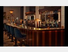 Elegante Bar mit einer geschwungenen Holztresenfläche, darauf Kerzen in Gläsern und Zapfanlagen. Im Hintergrund eine gut bestückte Rückwand-Bar mit hängenden Gläsern, flankiert von Kachelsäulen; davor stehen gepolsterte Barhocker mit blauem Stoffbezug.