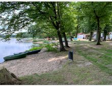 Sandstrand an einem See mit mehreren Ruderbooten am Ufer, umgeben von Bäumen. Im Hintergrund stehen Gebäude, darunter ein kleines weißes Haus mit einem roten Dach, und Sitzbereiche sind sichtbar.