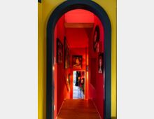 Schmaler, roter Flur mit Holzfußboden und farbenfrohen Gemälden an den Wänden, der durch einen gelb umrahmten Bogen betreten wird. Im Hintergrund ist ein weiterer Raum mit Möbeln, Pflanzen und einem Fenster erkennbar.
