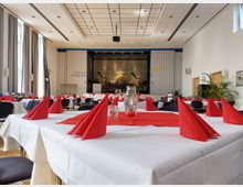 Ein Veranstaltungsraum mit gedeckten Tischen, weißen Tischdecken, roten Servietten und Glasflaschen in der Tischmitte. Im Hintergrund ist ein Ausstellungsflugzeug vor einer Wand mit der Aufschrift "Flugplatz Schönhagen" sichtbar.