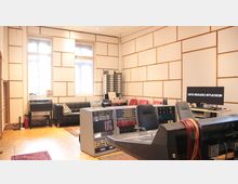 Ein Tonstudio mit akustisch gedämmten Wänden, einem Mischpult mit Kabeln, mehreren Audiogeräten und einem Computerbildschirm mit der Aufschrift „UFO Sound Studios“. Im Raum befinden sich außerdem eine schwarze Ledercouch, rote Stühle und Parkettboden.