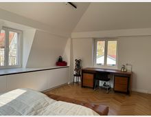 Dachgeschoss-Schlafzimmer mit Holzboden, einem Bett im Vordergrund, einer weißen Einbauschrankwand links und einem Schreibtisch aus Holz vor zwei Fenstern. Skulptur und kleine Dekorationen stehen auf Schreibtisch und Schrank.