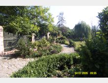 Gartenanlage mit einem geschwungenen, gepflasterten Weg, umgeben von Blumenbeeten und Büschen; links ein Zaun aus Holz und Ziegelpfeilern, im Hintergrund Bäume und eine weite Rasenfläche.