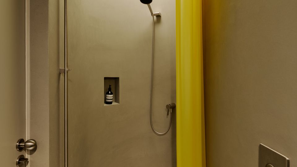 Kleine, moderne Dusche mit abgerundeten Wänden, einem runden Oberlicht und einem halbrunden, gesprenkelten Duschboden mit Ablauf. An der Wand sind eine Handbrause, eine eingelassene Ablagenische und ein gelber Duschvorhang zu sehen.