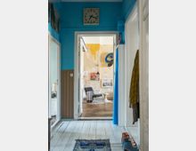 Ein Flur mit blau gestrichenen Wänden und weißem Holzboden, der zu einem Raum mit abgenutzter beigefarbener Wand führt, in dem ein schwarzer Stuhl, ein Beistelltisch und ein Wandbild stehen; ein blauer Vorhang grenzt den Bereich ab. Über der Tür hängen eine quadratische Uhr und Dekorationen, während Schuhe und ein Schal am rechten Bildrand sichtbar sind.