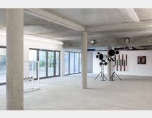 Großer, heller, moderner Ausstellungsraum mit Sichtbetondecke und -säulen, bodentiefen Fenstern und minimalistischem Design. Im Raum befinden sich Installationen, darunter eine Skulptur aus Metallrohren und ein Kunstwerk an der weißen Wand mit rosa und schwarzen Elementen.