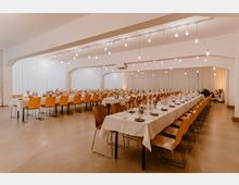 Großer, heller Saal mit langen, festlich gedeckten Banketttischen und gelben Holzstühlen. Von der Decke hängen einfache Glühbirnen, und der Raum ist von weißen Vorhängen umgeben; im Hintergrund stehen Pflanzen als Dekoration.