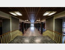 Großer Innenraum mit einer breiten Treppe aus schwarzem Marmor im Vordergrund, gelbem Geländer aus Metall, Säulenreihen auf beiden Seiten und einem gemusterten Holzdeckenpaneel. Am Ende des Raumes befindet sich eine Glastür, die nach draußen führt.